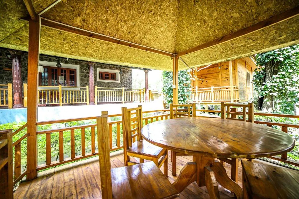A round wooden table in the dining gazebo, with the Stone Villa and the Wooden Cottage behind it among pepper vines