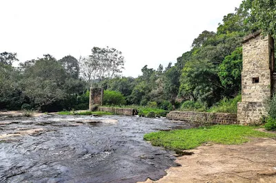 A shallow river running over flat rock past old stone towers, edged with forest